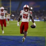 Ein Football-Spieler in einem rot-weißen Trikot rennt mit dem Ball in die Endzone, während seine Teamkollegen im Hintergrund feiern - ein elektrisierender Moment, der Emmett Johnsons Spielerprofil vor dem NFL Draft 2026 prägen könnte. Alt-Text wurde KI-generiert.