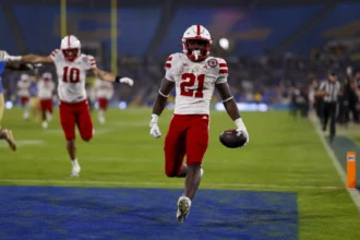 Ein Football-Spieler in einem rot-weißen Trikot rennt mit dem Ball in die Endzone, während seine Teamkollegen im Hintergrund feiern - ein elektrisierender Moment, der Emmett Johnsons Spielerprofil vor dem NFL Draft 2026 prägen könnte. Alt-Text wurde KI-generiert.