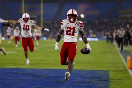 Ein Football-Spieler in einem rot-weißen Trikot rennt mit dem Ball in die Endzone, während seine Teamkollegen im Hintergrund feiern - ein elektrisierender Moment, der Emmett Johnsons Spielerprofil vor dem NFL Draft 2026 prägen könnte. Alt-Text wurde KI-generiert.