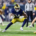 Ein Footballspieler in einem blau-goldenen Trikot, das an das Spielerprofil von Jadarian Price erinnert, rennt mit dem Ball, während Spieler in weißen Uniformen ihn verfolgen - und zeigt dabei Fähigkeiten, die im NFL Draft 2026 glänzen könnten. Alt-Text wurde KI-generiert.