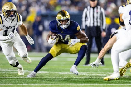 Ein Footballspieler in einem blau-goldenen Trikot, das an das Spielerprofil von Jadarian Price erinnert, rennt mit dem Ball, während Spieler in weißen Uniformen ihn verfolgen - und zeigt dabei Fähigkeiten, die im NFL Draft 2026 glänzen könnten. Alt-Text wurde KI-generiert.