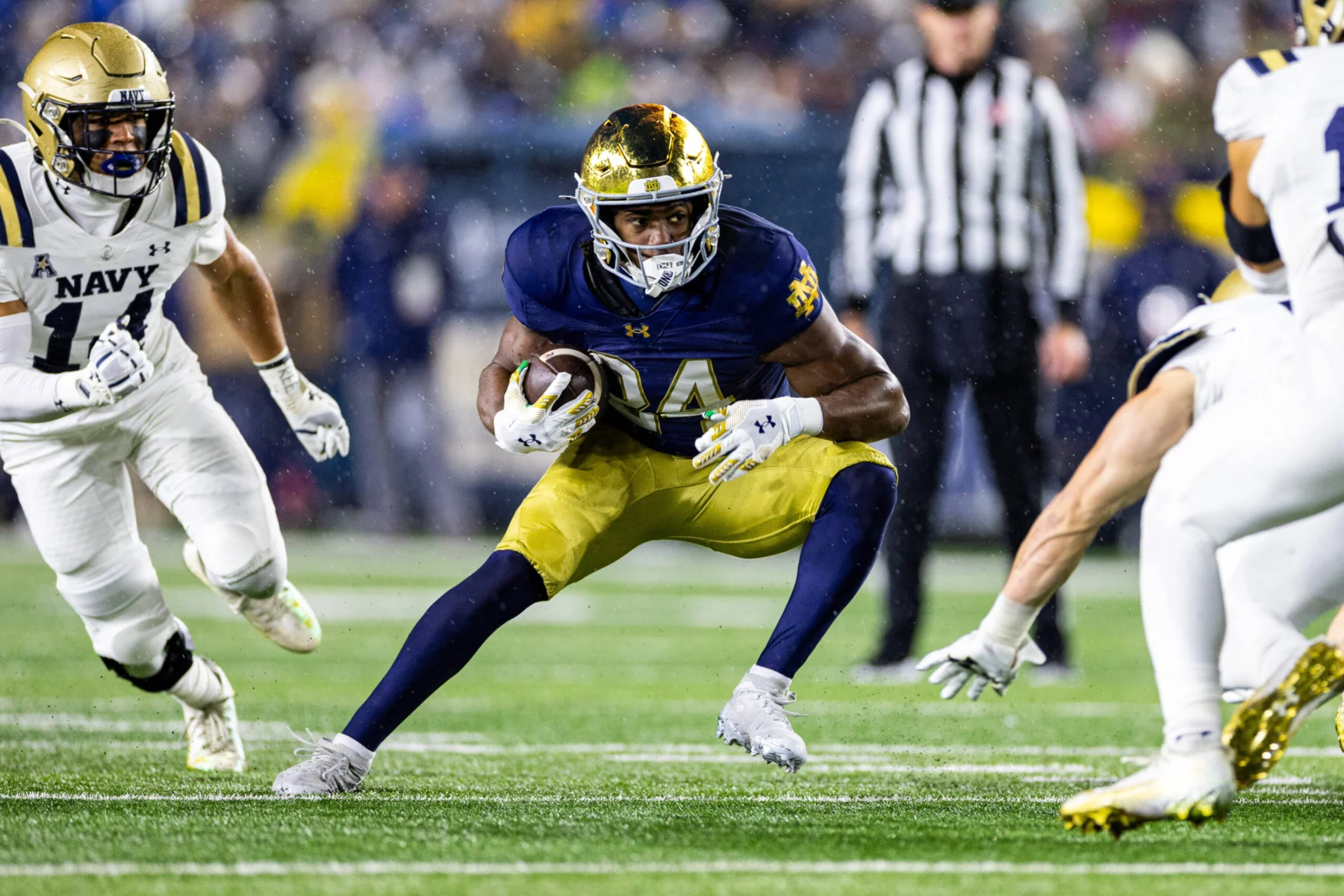 Ein Footballspieler in einem blau-goldenen Trikot, das an das Spielerprofil von Jadarian Price erinnert, rennt mit dem Ball, während Spieler in weißen Uniformen ihn verfolgen - und zeigt dabei Fähigkeiten, die im NFL Draft 2026 glänzen könnten. Alt-Text wurde KI-generiert.