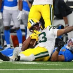 Dontayvion Wicks von den Packers liegt auf dem Feld und hält einen Football in der Hand, während ein Spieler der Detroit Lions während eines Footballspiels neben ihm auf dem Boden liegt. Alt-Text wurde KI-generiert.