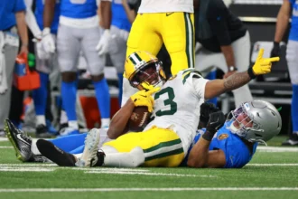 Dontayvion Wicks von den Packers liegt auf dem Feld und hält einen Football in der Hand, während ein Spieler der Detroit Lions während eines Footballspiels neben ihm auf dem Boden liegt. Alt-Text wurde KI-generiert.
