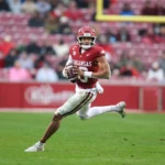 Der Arkansas-Football-Spieler Taylen Green in einem roten Trikot läuft während eines Spiels mit dem Ball auf einer Wiese und stellt seine Fähigkeiten für den NFL Draft 2026 unter Beweis, während im Hintergrund Stadionsitze zu sehen sind. Alt-Text wurde KI-generiert.