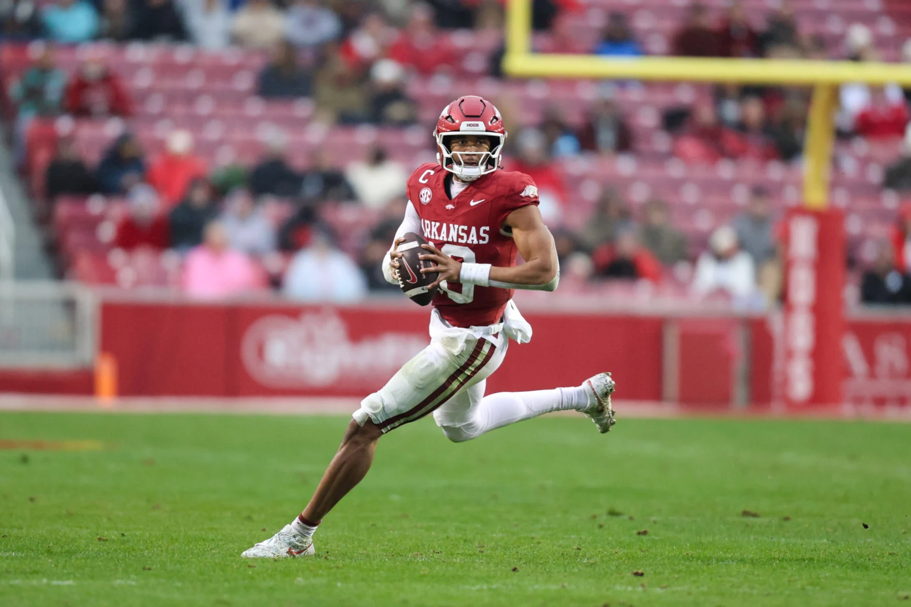 Der Arkansas-Football-Spieler Taylen Green in einem roten Trikot läuft während eines Spiels mit dem Ball auf einer Wiese und stellt seine Fähigkeiten für den NFL Draft 2026 unter Beweis, während im Hintergrund Stadionsitze zu sehen sind. Alt-Text wurde KI-generiert.