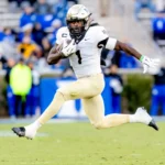 NFL Draft 2026 Kandidat Demond Claiborne zeigt seine Fähigkeiten als Footballspieler in einem weiß-goldenen Trikot, indem er in der Luft springt, während er mit dem Ball läuft - ein beeindruckender Moment für sein Spielerprofil. Alt-Text wurde KI-generiert.