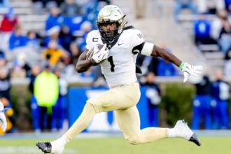 NFL Draft 2026 Kandidat Demond Claiborne zeigt seine Fähigkeiten als Footballspieler in einem weiß-goldenen Trikot, indem er in der Luft springt, während er mit dem Ball läuft - ein beeindruckender Moment für sein Spielerprofil. Alt-Text wurde KI-generiert.