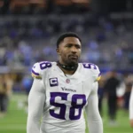 Jonathan Greenard, der ein weißes Minnesota Vikings-Trikot mit der Nummer 58 trägt, steht auf dem Spielfeld und blickt nach vorne, während im Hintergrund weitere Personen zu sehen sind. Der entschlossene Wikinger beobachtet die Szene, während das Spiel gegen die Eagles immer intensiver wird. Alt-Text wurde KI-generiert.