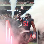 Will Anderson, der für die Texans die Nummer 51 trägt, betritt das Spielfeld durch Rauch und Lichter und hebt seinen rechten Arm, während die Fans auf der Tribüne zusehen. Alt-Text wurde KI-generiert.