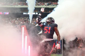 Will Anderson, der für die Texans die Nummer 51 trägt, betritt das Spielfeld durch Rauch und Lichter und hebt seinen rechten Arm, während die Fans auf der Tribüne zusehen. Alt-Text wurde KI-generiert.