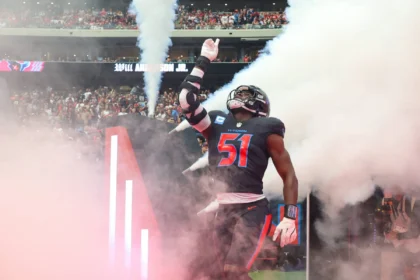 Will Anderson, der für die Texans die Nummer 51 trägt, betritt das Spielfeld durch Rauch und Lichter und hebt seinen rechten Arm, während die Fans auf der Tribüne zusehen. Alt-Text wurde KI-generiert.