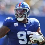 Dexter Lawrence, im blauen Trikot und Helm der New York Giants mit der Nummer 97, hält einen Football in der Hand, während er während eines Spiels auf das Spielfeld rennt - vielleicht zum Feiern nach seiner Vertragsverlängerung oder gegen Rivalen wie die Bengals. Alt-Text wurde KI-generiert.