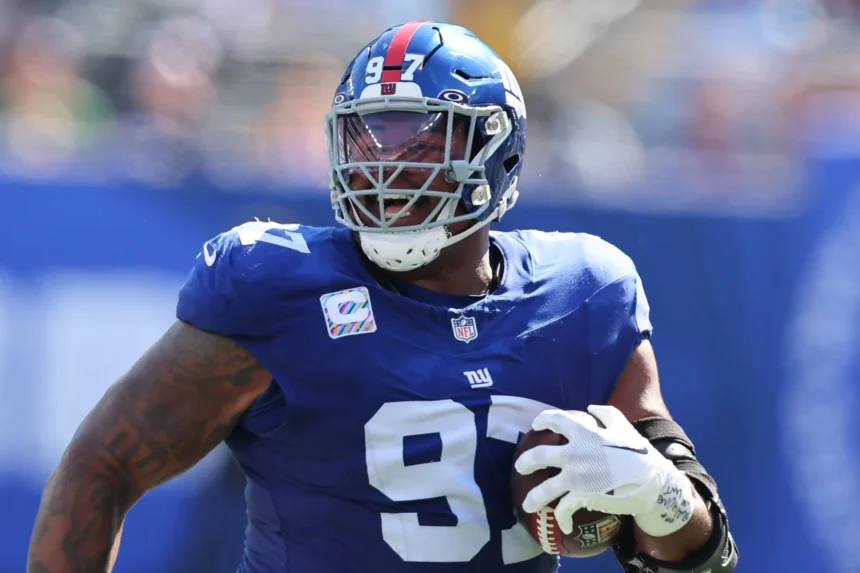 Dexter Lawrence, im blauen Trikot und Helm der New York Giants mit der Nummer 97, hält einen Football in der Hand, während er während eines Spiels auf das Spielfeld rennt - vielleicht zum Feiern nach seiner Vertragsverlängerung oder gegen Rivalen wie die Bengals. Alt-Text wurde KI-generiert.