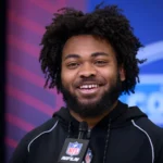 Ein Mann mit lockigem Haar und Bart, der einen schwarzen NFL-Kapuzenpulli trägt, lächelt, während er bei einer Veranstaltung mit einem NFL Draft 2026-Logo im Hintergrund in ein Mikrofon spricht und möglicherweise über das Spielerprofil von Desmond Reid spricht. Alt-Text wurde KI-generiert.