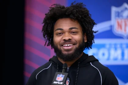 Ein Mann mit lockigem Haar und Bart, der einen schwarzen NFL-Kapuzenpulli trägt, lächelt, während er bei einer Veranstaltung mit einem NFL Draft 2026-Logo im Hintergrund in ein Mikrofon spricht und möglicherweise über das Spielerprofil von Desmond Reid spricht. Alt-Text wurde KI-generiert.
