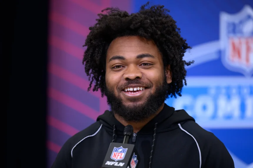 Ein Mann mit lockigem Haar und Bart, der einen schwarzen NFL-Kapuzenpulli trägt, lächelt, während er bei einer Veranstaltung mit einem NFL Draft 2026-Logo im Hintergrund in ein Mikrofon spricht und möglicherweise über das Spielerprofil von Desmond Reid spricht. Alt-Text wurde KI-generiert.