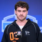 Eine Person, die einen schwarzen Kapuzenpulli mit der Aufschrift "02" trägt, spricht während einer Presseveranstaltung vor einem NFL-Mikrofon, mit Microsoft Copilot-Branding im Hintergrund, über den bevorstehenden NFL Draft 2026 und aufstrebende Talente wie Luke Altmyer. Alt-Text wurde KI-generiert.