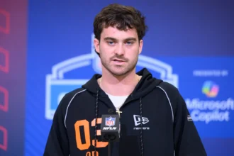 Eine Person, die einen schwarzen Kapuzenpulli mit der Aufschrift "02" trägt, spricht während einer Presseveranstaltung vor einem NFL-Mikrofon, mit Microsoft Copilot-Branding im Hintergrund, über den bevorstehenden NFL Draft 2026 und aufstrebende Talente wie Luke Altmyer. Alt-Text wurde KI-generiert.