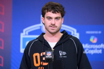 Eine Person, die einen schwarzen Kapuzenpulli mit der Aufschrift "02" trägt, spricht während einer Presseveranstaltung vor einem NFL-Mikrofon, mit Microsoft Copilot-Branding im Hintergrund, über den bevorstehenden NFL Draft 2026 und aufstrebende Talente wie Luke Altmyer. Alt-Text wurde KI-generiert.