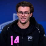 Ein Mann mit Brille und dunklem Haar, der einen schwarzen Kapuzenpulli mit der Nummer 14 in Rosa trägt, spricht während des NFL Draft 2026 in ein Mikrofon vor blauem Hintergrund. Alt-Text wurde KI-generiert.