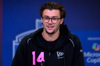 Ein Mann mit Brille und dunklem Haar, der einen schwarzen Kapuzenpulli mit der Nummer 14 in Rosa trägt, spricht während des NFL Draft 2026 in ein Mikrofon vor blauem Hintergrund. Alt-Text wurde KI-generiert.