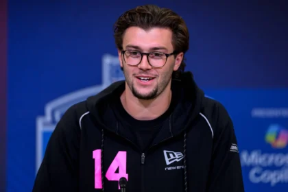 Ein Mann mit Brille und dunklem Haar, der einen schwarzen Kapuzenpulli mit der Nummer 14 in Rosa trägt, spricht während des NFL Draft 2026 in ein Mikrofon vor blauem Hintergrund. Alt-Text wurde KI-generiert.