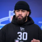 Ein Mann mit Bart und schwarzer Mütze, der einen schwarzen Kapuzenpulli mit der Nummer 30 trägt, steht bei einer Veranstaltung vor einer Kulisse für den NFL Draft 2026. Alt-Text wurde KI-generiert.