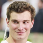 Ein junger Mann mit kurzen braunen Haaren, der ein weißes Hemd und einen Ohrhörer trägt, schaut leicht zur Seite und lächelt. Der Hintergrund ist unscharf und deutet auf Fernando Mendozas Begeisterung für den NFL Draft 2026 und seine Träume mit den Raiders hin. Alt-Text wurde KI-generiert.