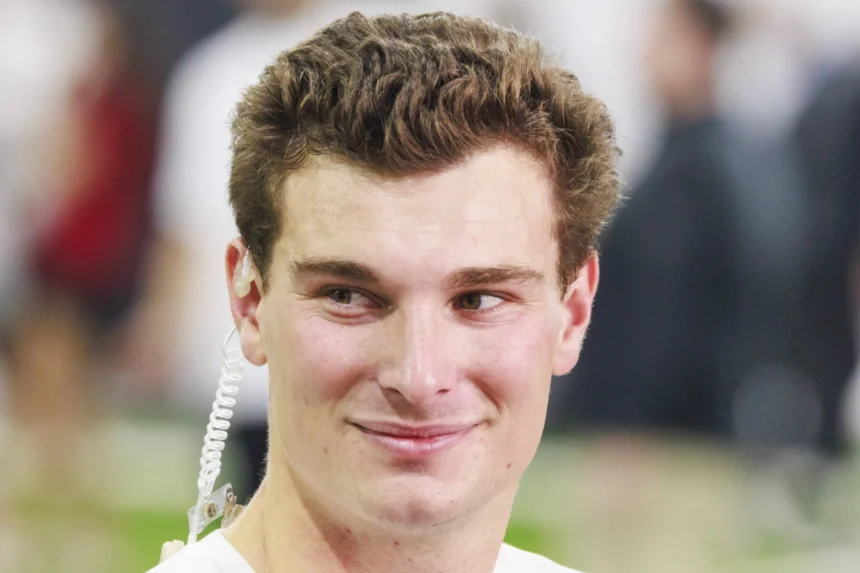 Ein junger Mann mit kurzen braunen Haaren, der ein weißes Hemd und einen Ohrhörer trägt, schaut leicht zur Seite und lächelt. Der Hintergrund ist unscharf und deutet auf Fernando Mendozas Begeisterung für den NFL Draft 2026 und seine Träume mit den Raiders hin. Alt-Text wurde KI-generiert.