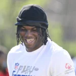 Eine Person, die eine schwarze Kopfbedeckung und ein weißes NFL-Trikot mit einem Special Olympics-Logo trägt, lächelt an einem sonnigen Tag im Freien und inspiriert künftige Edge Rusher im Vorfeld des NFL Draft 2026. Alt-Text wurde KI-generiert.