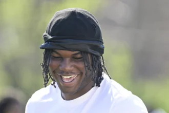 Eine Person, die eine schwarze Kopfbedeckung und ein weißes NFL-Trikot mit einem Special Olympics-Logo trägt, lächelt an einem sonnigen Tag im Freien und inspiriert künftige Edge Rusher im Vorfeld des NFL Draft 2026. Alt-Text wurde KI-generiert.