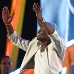 Ein Mann in einer hellen Jacke und einer blauen Mütze hebt lächelnd die Arme auf einer hell erleuchteten Bühne mit bunten Lichtern im Hintergrund und feiert etwas, das ein Draft-Event zu sein scheint, möglicherweise für die Titans oder mit Carnell Tate. Alt-Text wurde KI-generiert.