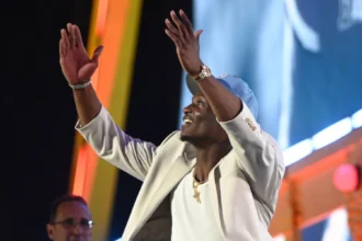 Ein Mann in einer hellen Jacke und einer blauen Mütze hebt lächelnd die Arme auf einer hell erleuchteten Bühne mit bunten Lichtern im Hintergrund und feiert etwas, das ein Draft-Event zu sein scheint, möglicherweise für die Titans oder mit Carnell Tate. Alt-Text wurde KI-generiert.