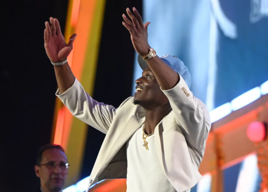 Ein Mann in einer hellen Jacke und einer blauen Mütze hebt lächelnd die Arme auf einer hell erleuchteten Bühne mit bunten Lichtern im Hintergrund und feiert etwas, das ein Draft-Event zu sein scheint, möglicherweise für die Titans oder mit Carnell Tate. Alt-Text wurde KI-generiert.