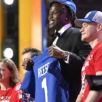 Ein Footballspieler im Anzug und mit blauer Mütze, der als Arvell Reese identifiziert wird, hält ein blaues Trikot mit der Aufschrift "REESE 1" in der Hand, während er mit drei lächelnden Menschen auf der Bühne einer Veranstaltung steht. Alt-Text wurde KI-generiert.