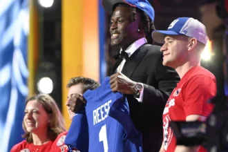 Ein Footballspieler im Anzug und mit blauer Mütze, der als Arvell Reese identifiziert wird, hält ein blaues Trikot mit der Aufschrift "REESE 1" in der Hand, während er mit drei lächelnden Menschen auf der Bühne einer Veranstaltung steht. Alt-Text wurde KI-generiert.