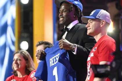 Ein Footballspieler im Anzug und mit blauer Mütze, der als Arvell Reese identifiziert wird, hält ein blaues Trikot mit der Aufschrift "REESE 1" in der Hand, während er mit drei lächelnden Menschen auf der Bühne einer Veranstaltung steht. Alt-Text wurde KI-generiert.