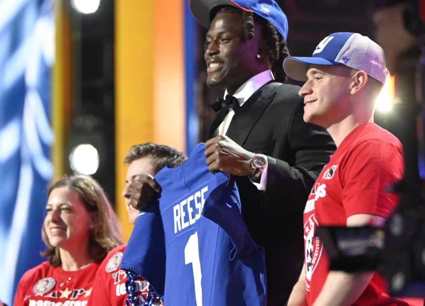 Ein Footballspieler im Anzug und mit blauer Mütze, der als Arvell Reese identifiziert wird, hält ein blaues Trikot mit der Aufschrift "REESE 1" in der Hand, während er mit drei lächelnden Menschen auf der Bühne einer Veranstaltung steht. Alt-Text wurde KI-generiert.