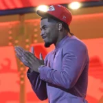 Mansoor Delane, ein Cornerback, faltet seine Hände zusammen, während er eine rote Kansas City Chiefs-Mütze und einen lilafarbenen Anzug trägt und vor einem leuchtend roten und orangen Hintergrund steht. Alt-Text wurde KI-generiert.