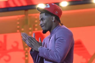 Mansoor Delane, ein Cornerback, faltet seine Hände zusammen, während er eine rote Kansas City Chiefs-Mütze und einen lilafarbenen Anzug trägt und vor einem leuchtend roten und orangen Hintergrund steht. Alt-Text wurde KI-generiert.