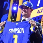 Ein lächelnder Mann im Anzug und mit Teamkappe hält ein blaues Rams-Football-Trikot mit der Aufschrift "Simpson" und der Nummer 1 hoch und feiert damit Ty Simpson als potenziellen Stafford-Nachfolger bei einer Draft-Veranstaltung. Alt-Text wurde KI-generiert.