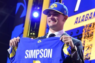 Ein lächelnder Mann im Anzug und mit Teamkappe hält ein blaues Rams-Football-Trikot mit der Aufschrift "Simpson" und der Nummer 1 hoch und feiert damit Ty Simpson als potenziellen Stafford-Nachfolger bei einer Draft-Veranstaltung. Alt-Text wurde KI-generiert.