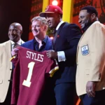 Vier Männer stehen auf der Bühne, einer von ihnen hält ein kastanienbraunes Football-Trikot mit der Aufschrift "STYLES 1". Zwei Männer tragen hellbraune Jacken mit Hall of Fame-Aufnähern, während der NFL Draft 2026 die Big Ten-Aussichten der Rams hervorhebt. Alt-Text wurde KI-generiert.