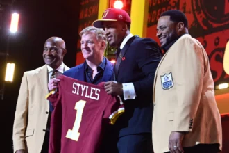 Vier Männer stehen auf der Bühne, einer von ihnen hält ein kastanienbraunes Football-Trikot mit der Aufschrift "STYLES 1". Zwei Männer tragen hellbraune Jacken mit Hall of Fame-Aufnähern, während der NFL Draft 2026 die Big Ten-Aussichten der Rams hervorhebt. Alt-Text wurde KI-generiert.