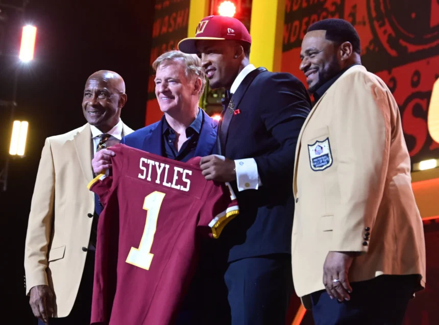 Vier Männer stehen auf der Bühne, einer von ihnen hält ein kastanienbraunes Football-Trikot mit der Aufschrift "STYLES 1". Zwei Männer tragen hellbraune Jacken mit Hall of Fame-Aufnähern, während der NFL Draft 2026 die Big Ten-Aussichten der Rams hervorhebt. Alt-Text wurde KI-generiert.