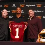 Drei Männer stehen vor einer Kulisse der Washington Commanders, einer von ihnen hält ein "STYLES 1"-Trikot - ein NFL Draft 2026-Gewinner-Moment - während auf dem Tisch daneben ein Helm der Commanders liegt. Alt-Text wurde KI-generiert.