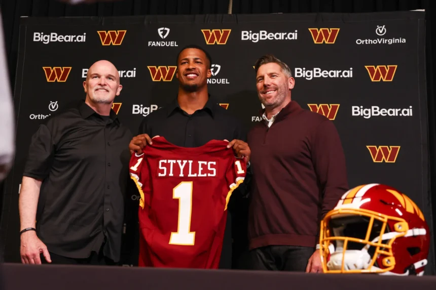 Drei Männer stehen vor einer Kulisse der Washington Commanders, einer von ihnen hält ein "STYLES 1"-Trikot - ein NFL Draft 2026-Gewinner-Moment - während auf dem Tisch daneben ein Helm der Commanders liegt. Alt-Text wurde KI-generiert.