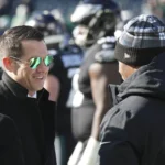 Ein Mann mit schwarzem Mantel und Sonnenbrille lächelt, während er mit einer anderen Person, die eine Wintermütze trägt, auf einem Footballfeld in New York spricht. Im Hintergrund sind Spieler zu sehen, die möglicherweise über die Verlängerung des Vertrags von GM Joe Schoen diskutieren. Alt-Text wurde KI-generiert.