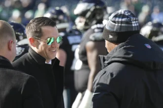 Ein Mann mit schwarzem Mantel und Sonnenbrille lächelt, während er mit einer anderen Person, die eine Wintermütze trägt, auf einem Footballfeld in New York spricht. Im Hintergrund sind Spieler zu sehen, die möglicherweise über die Verlängerung des Vertrags von GM Joe Schoen diskutieren. Alt-Text wurde KI-generiert.